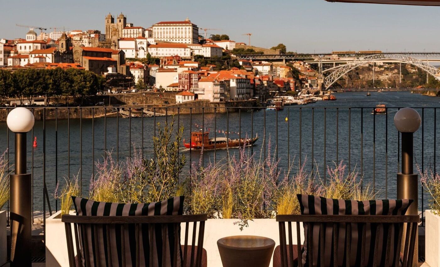 Two lounge chairs facing a scenic river with boats, a cityscape of colorful buildings and a bridge in the background. An umbrella provides shade on a sunny day.
