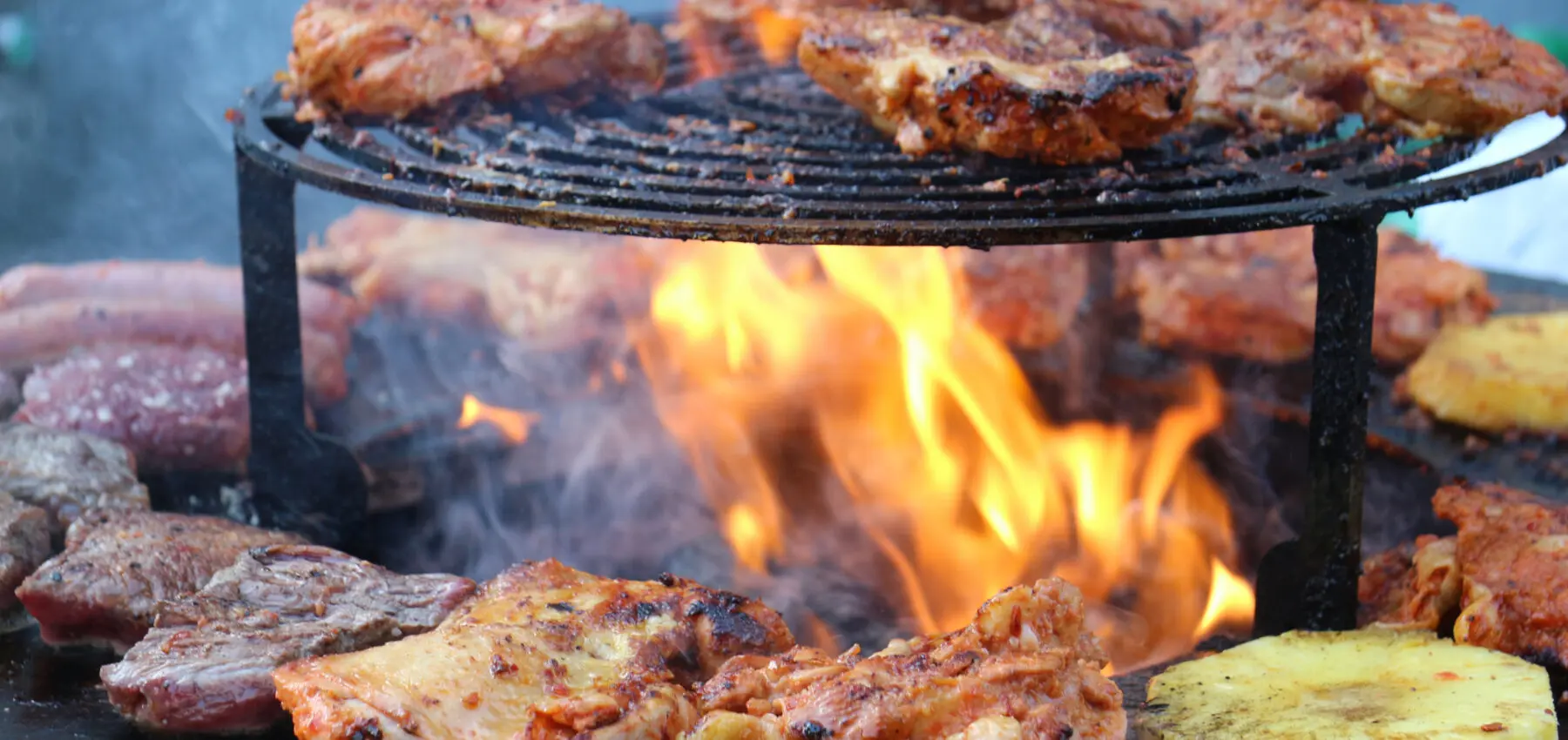 Grilled chicken, beef, and pineapple slices cook over an open flame on a round metal grill, with smoke rising from the heat.