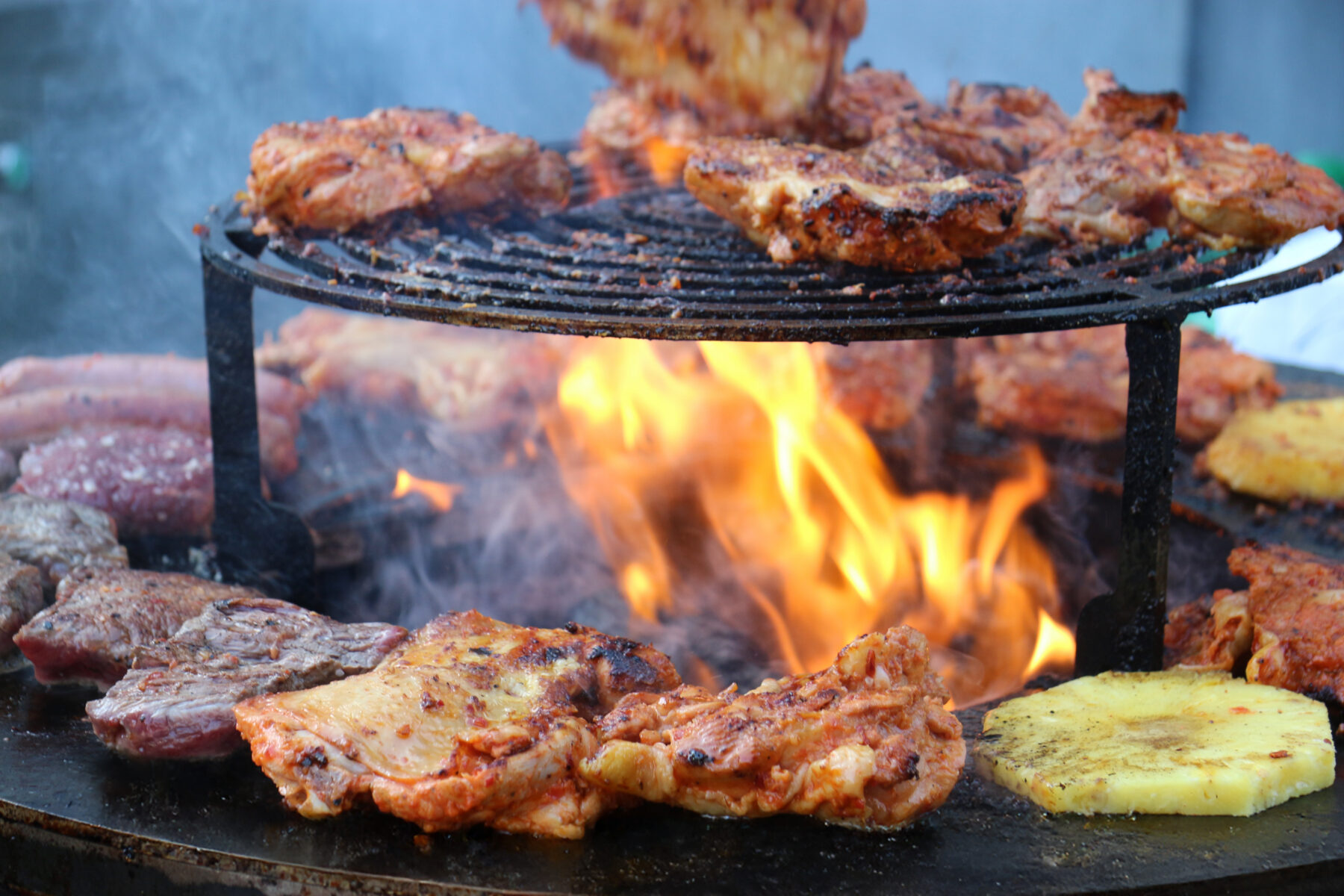 Grilled chicken, beef, and pineapple slices cook over an open flame on a round metal grill, with smoke rising from the heat.