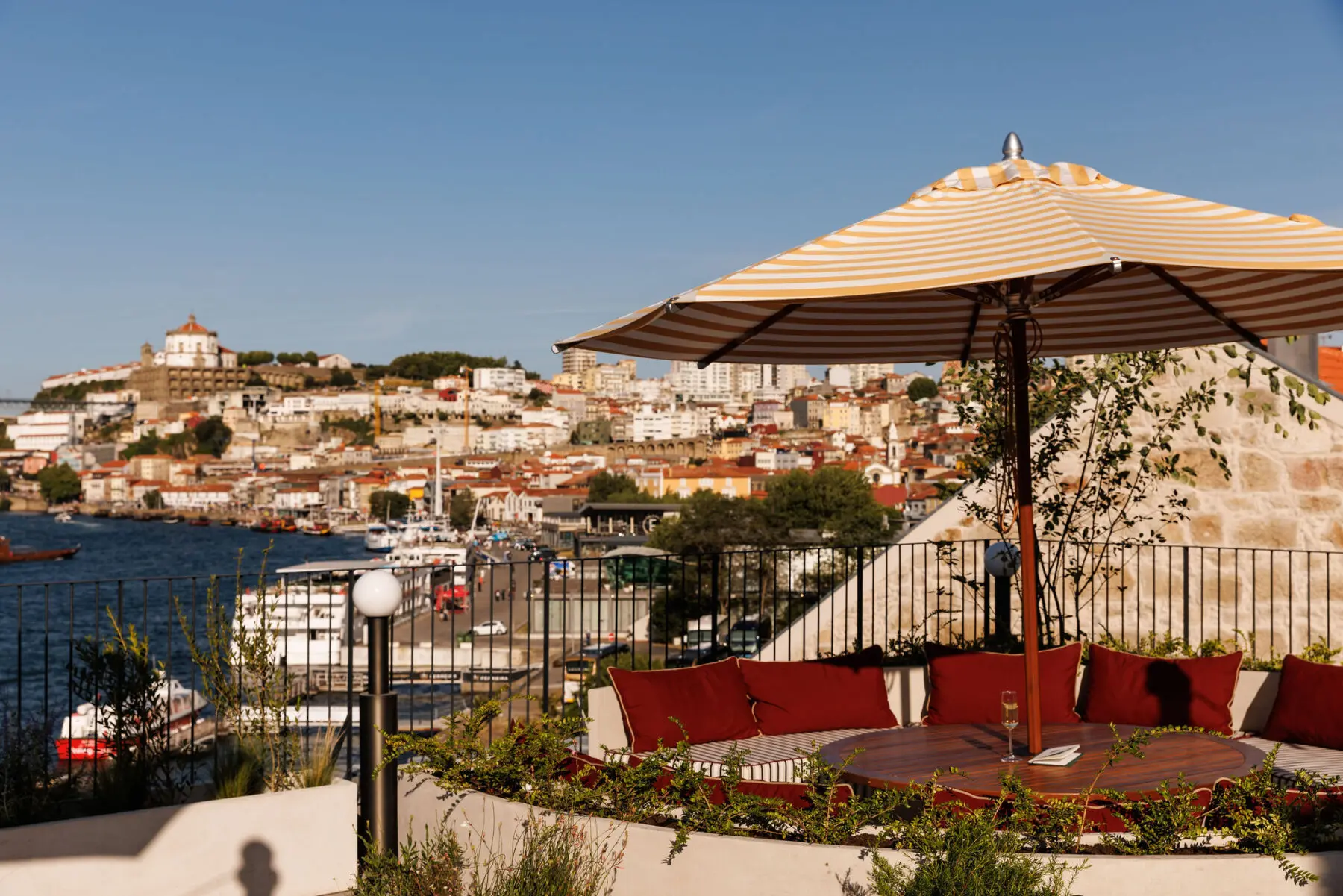 A sunlit outdoor seating area with red cushions and a striped umbrella overlooks a river, boats, and a Porto cityscape under a clear blue sky.