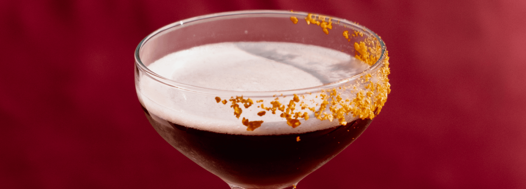A close-up of a cocktail in a coupe glass with a foamy top and a partial golden-brown sugar or spice rim, set against a deep red background.