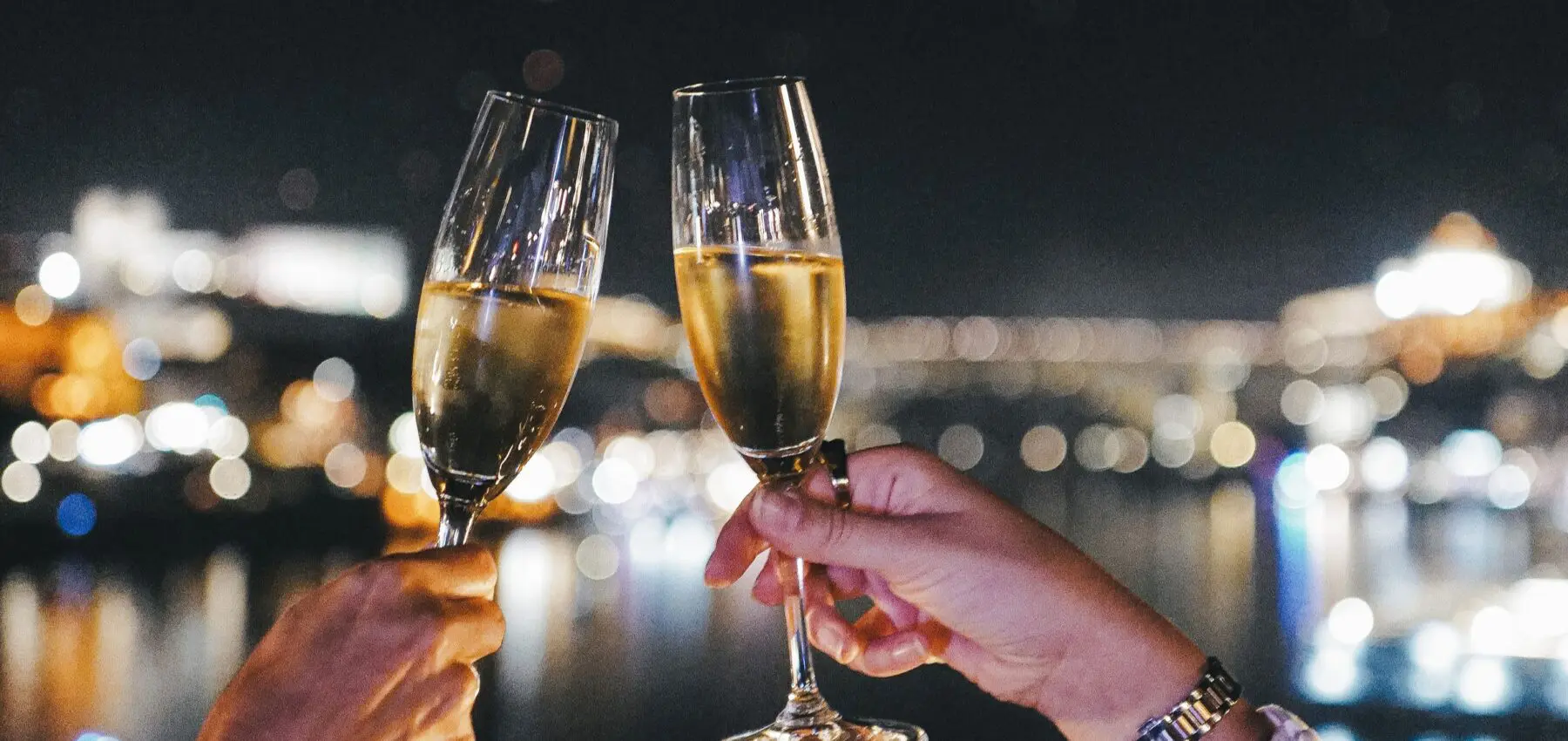 Deux mains s'entrechoquant dans des coupes de champagne la nuit, avec des lumières de ville floues et une rivière en arrière-plan, suggérant un moment de célébration ou de romantisme.
