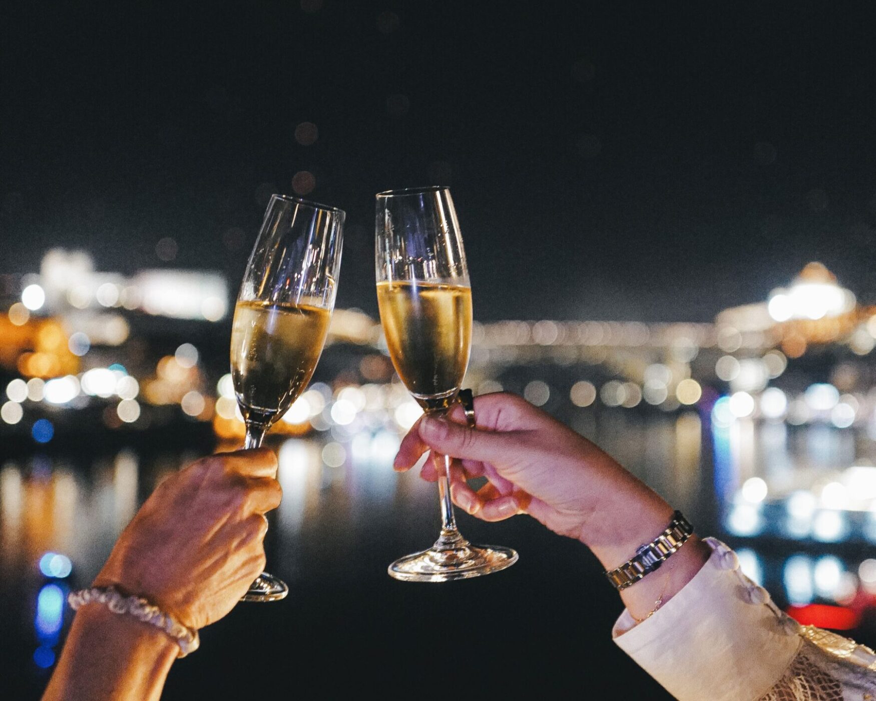 Deux mains s'entrechoquant dans des coupes de champagne la nuit, avec des lumières de ville floues et une rivière en arrière-plan, suggérant un moment de célébration ou de romantisme.