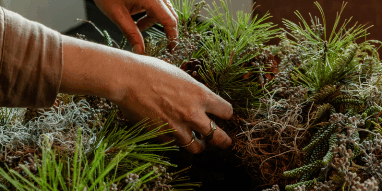 A person’s hands arranging pine branches, moss, and small cones, likely making a holiday wreath.