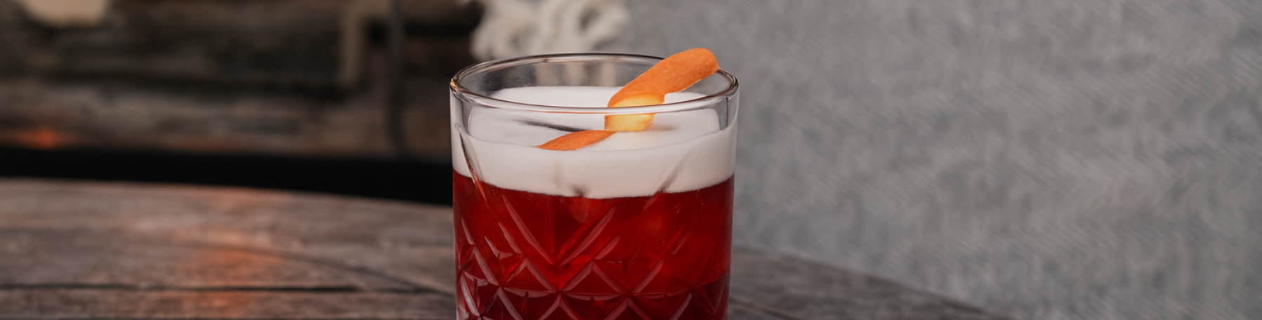 Un vaso de cóctel rojo con espuma blanca y un adorno de piel de naranja sobre una mesa de madera rústica.