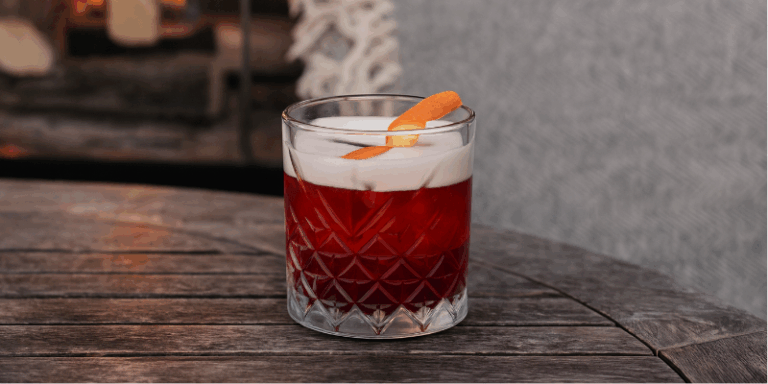 A red cocktail with a frothy white layer on top and an orange peel garnish, served in a patterned glass tumbler on a round wooden table.