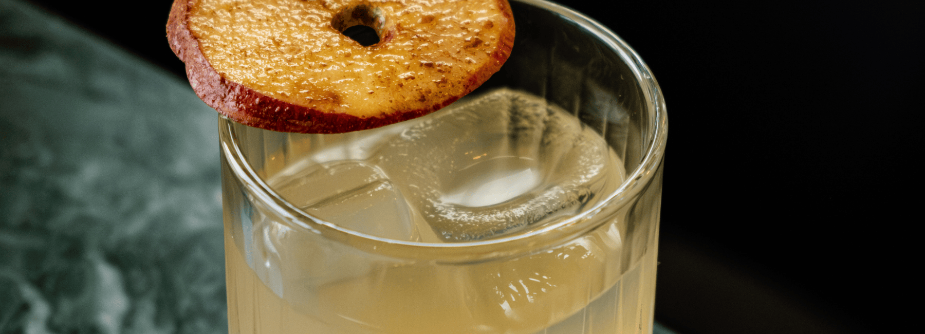 A glass of light-colored drink with large ice cubes, garnished with a dried apple slice on the rim, set on a green marble surface.