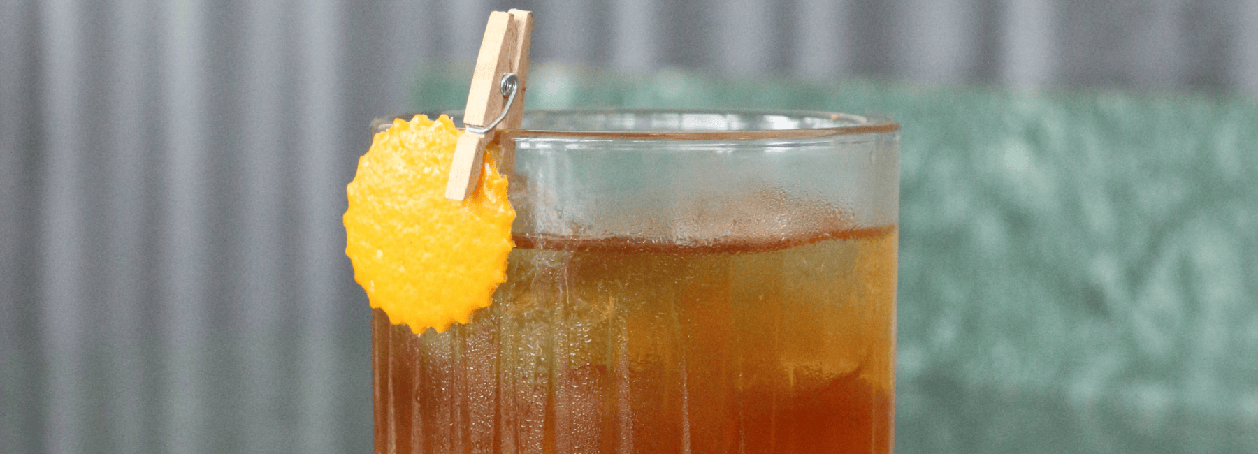 Primer plano de un vaso lleno de una bebida de color ámbar, adornado con una rodaja redonda de naranja sujeta al borde con una pequeña pinza de madera. El fondo está difuminado.