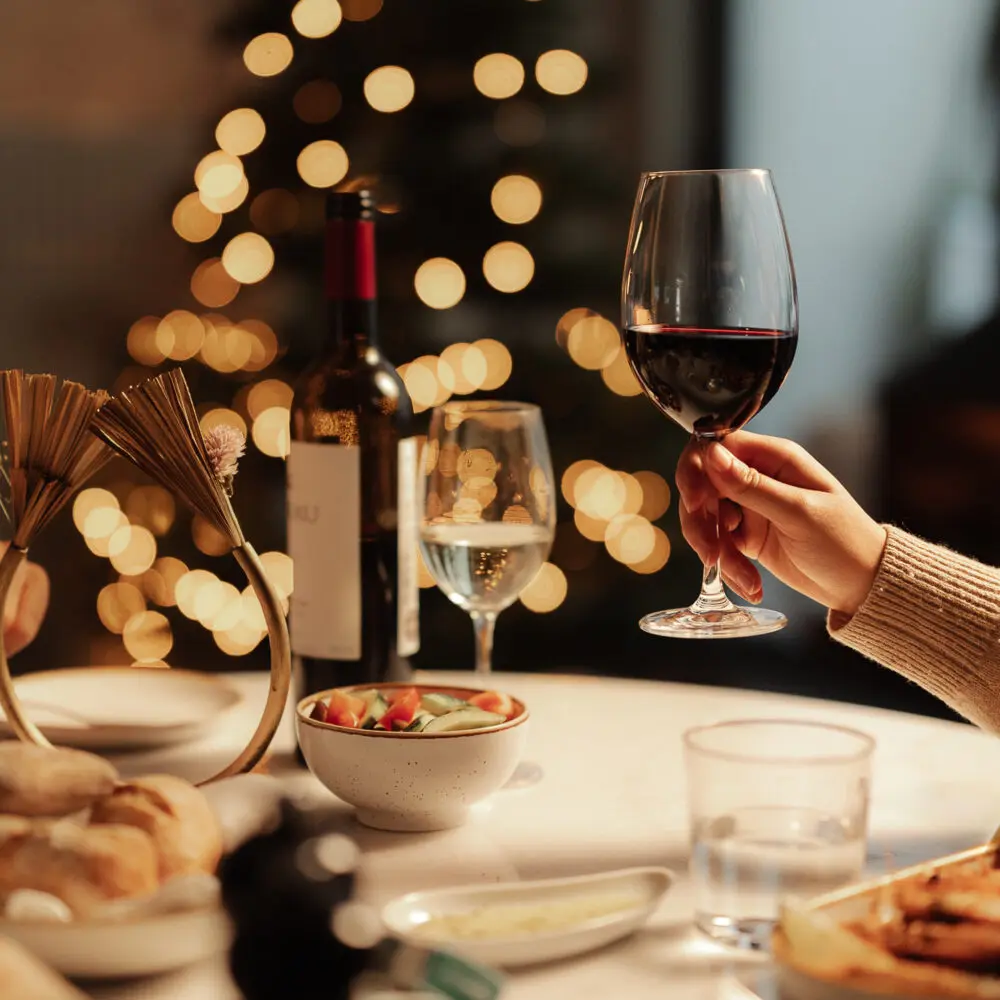 A hand holds a glass of red wine over a festive dinner table with bread, salad, and wine, warmly lit by a fireplace at Hotel Porto and blurred holiday lights in the background.