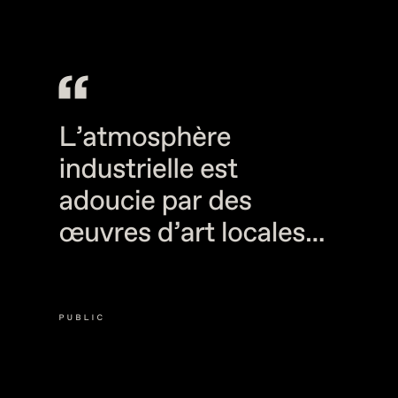 Le texte français blanc sur fond noir se lit comme suit : "L'atmosphère industrielle est adoucie par des œuvres d'art locales" : "L'atmosphère industrielle est adoucie par des œuvres d'art locales..." avec un petit "PUBLIC" en dessous.