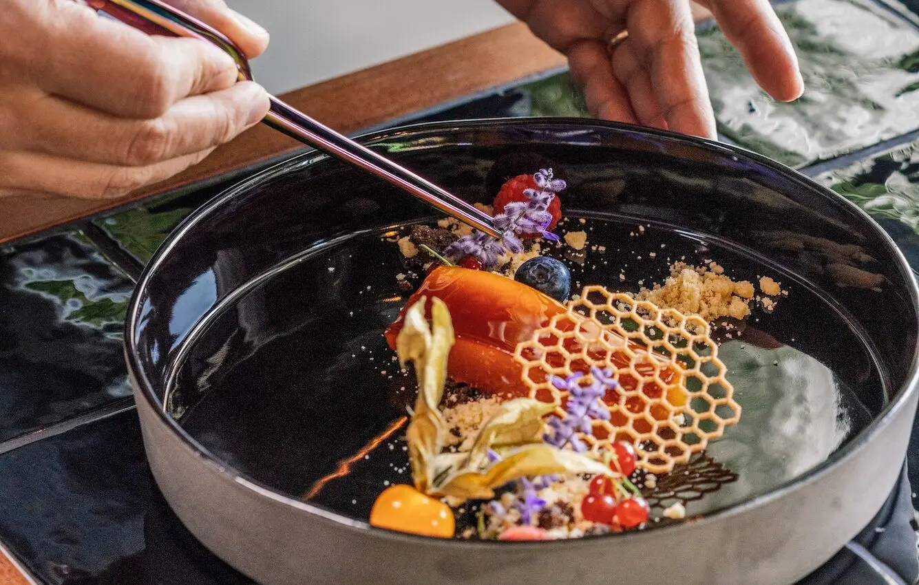 A restaurant chef arranges a colorful gourmet dessert with edible flowers, berries, a honeycomb-shaped garnish, and crumbs on a black plate using metal chopsticks.