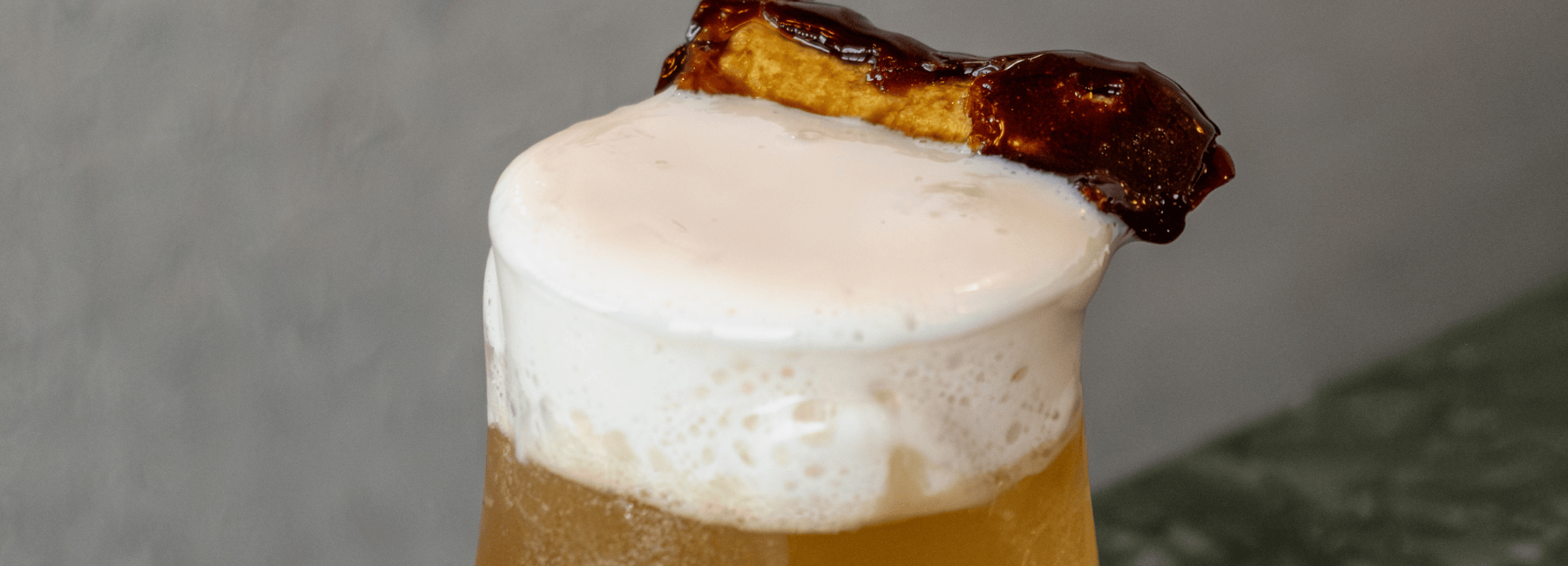 Gros plan sur un verre de bière moussant, surmonté d'une épaisse mousse et garni d'un morceau de bacon caramélisé reposant sur le bord.