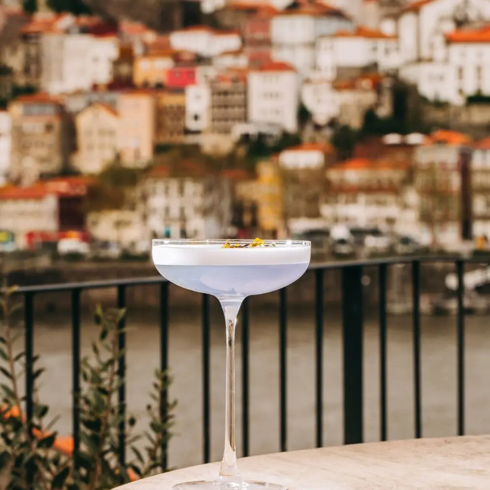 A cocktail glass with a light blue drink sits on a round table outdoors, crafted by a restaurant chef, against a blurred backdrop of colorful hillside buildings near a river.