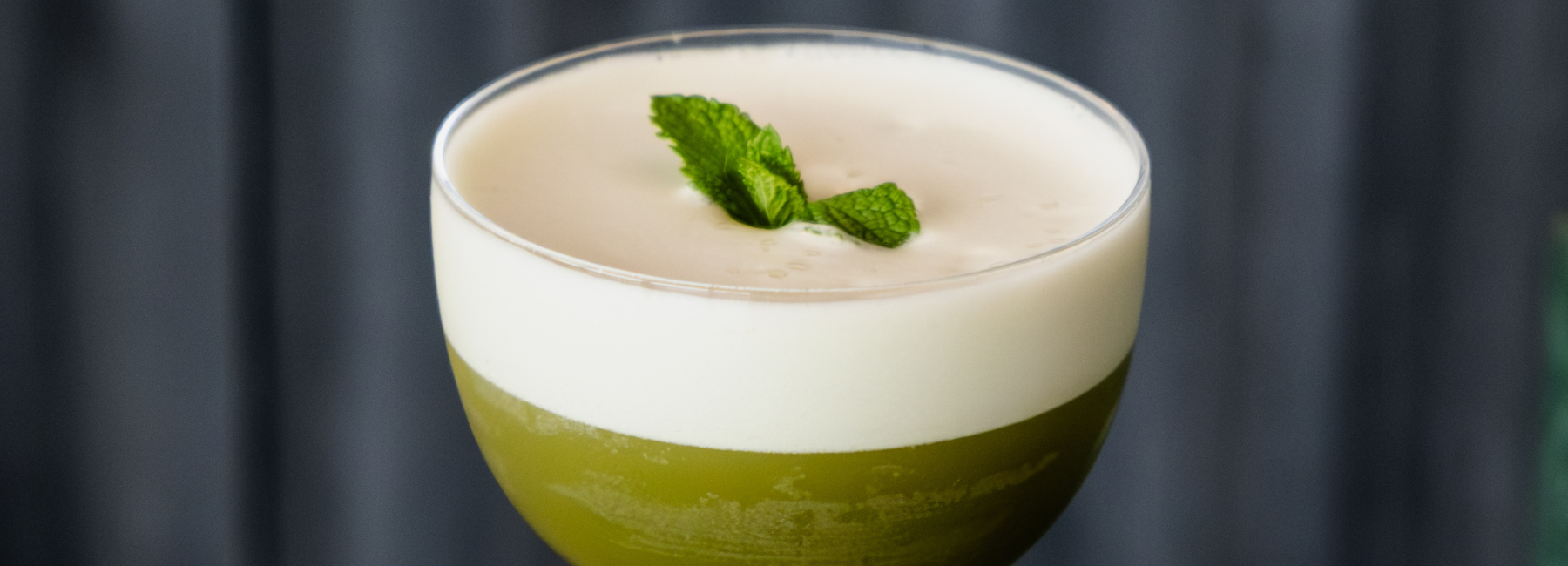 Un vaso lleno de una bebida verde coronada con una gruesa capa de espuma blanca, adornada con dos hojas de menta fresca, sobre un fondo oscuro y borroso.
