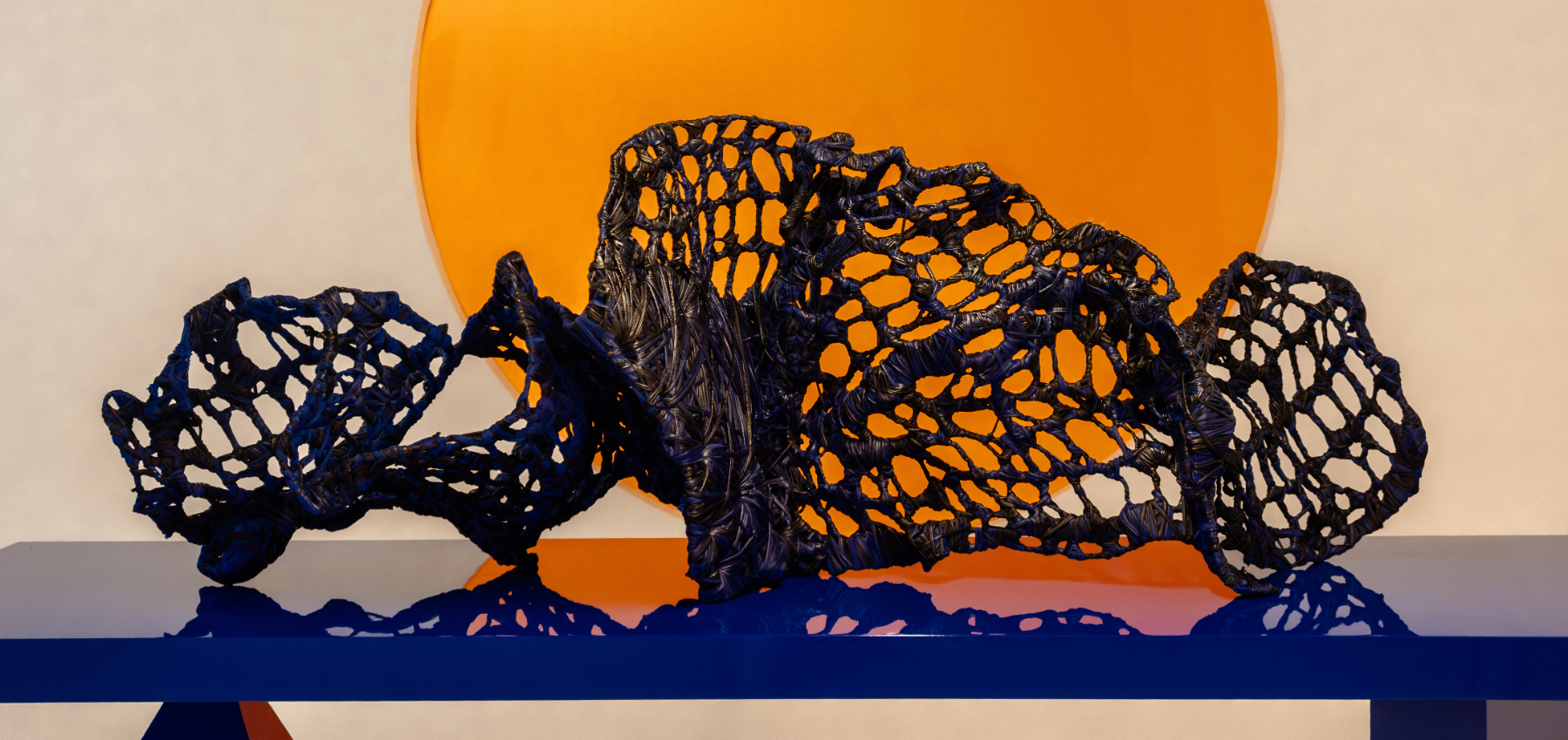 A dark, net-like abstract sculpture rests on a glossy blue surface, with a large orange circle in the background against a neutral wall.