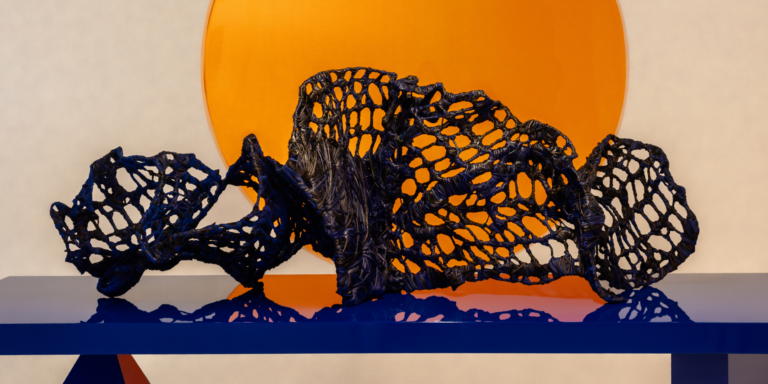 A dark, net-like abstract sculpture rests on a glossy blue surface, with a large orange circle in the background against a neutral wall.