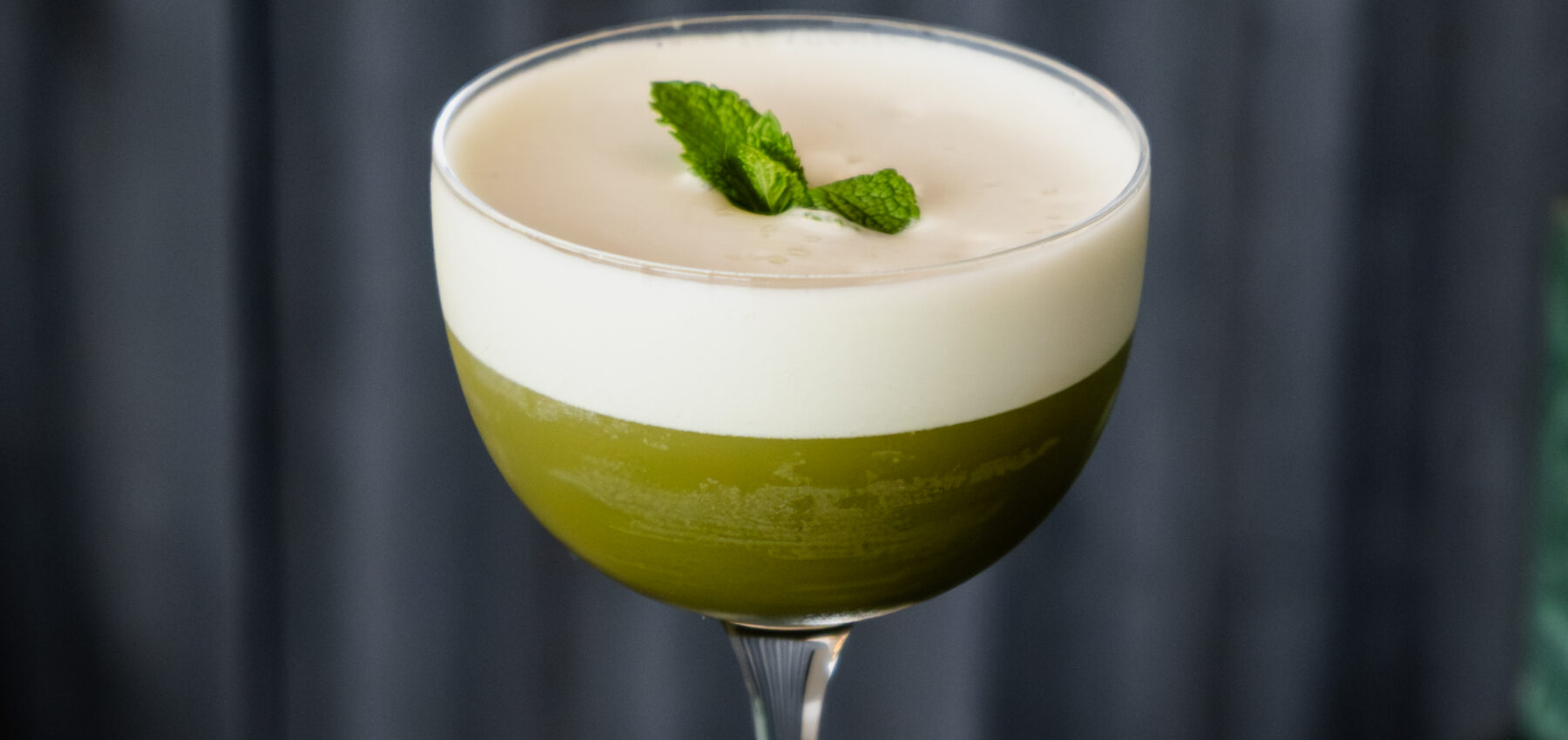 Un verre à coupe rempli d'un cocktail vert surmonté d'une épaisse couche de mousse blanche et garni d'un petit brin de menthe, le tout sur un fond sombre et texturé.