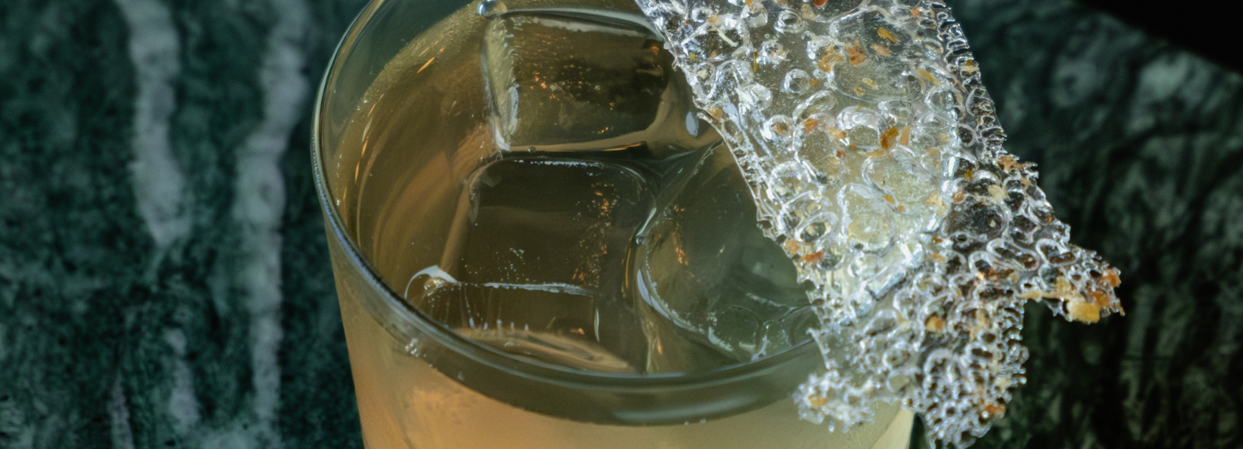 Gros plan sur un cocktail dans un verre avec des glaçons, garni d'un grand morceau texturé de nid d'abeille sur une surface marbrée verte.