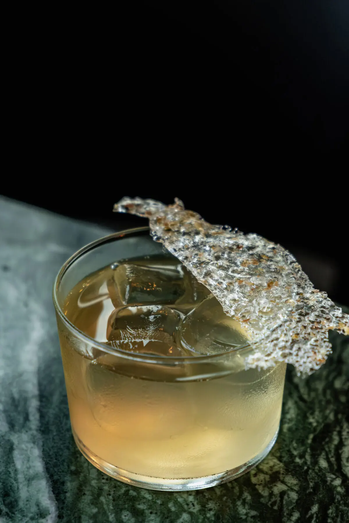 A cocktail served in a short glass with ice, topped with a decorative, translucent garnish, resting on a green marble surface against a dark background.