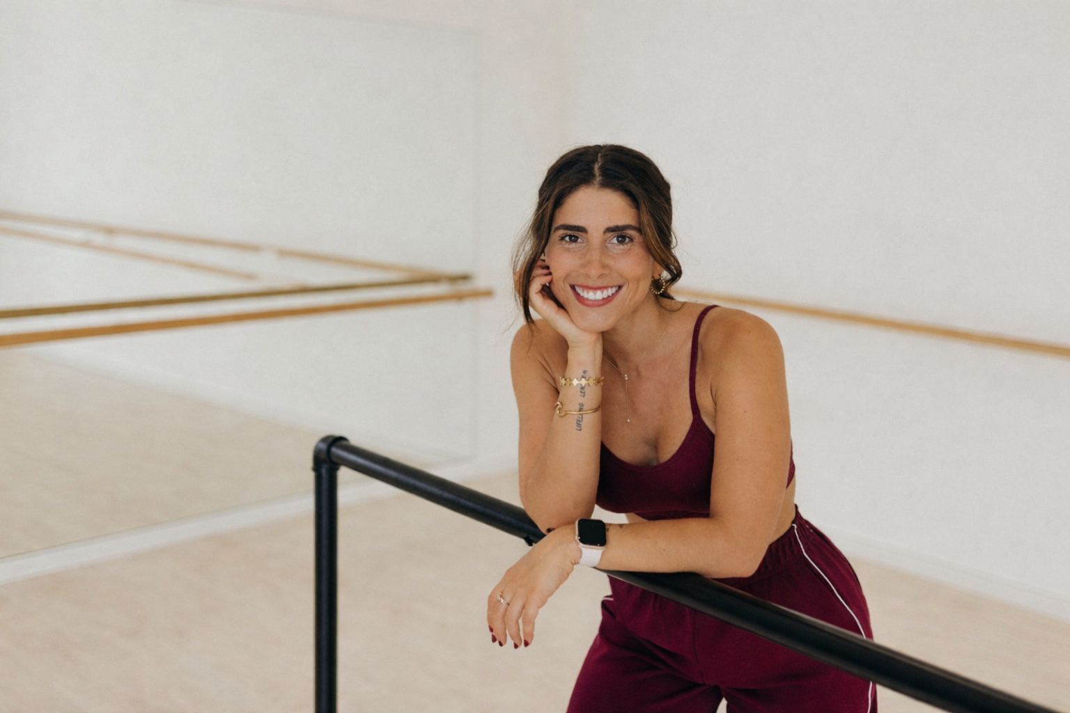 A smiling woman wearing a burgundy sleeveless jumpsuit leans on a black ballet barre in a bright studio with mirrors and wooden barres in the background.