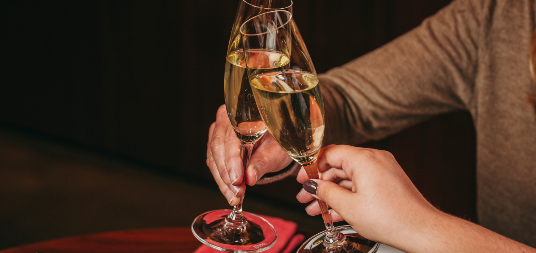 Dos personas chocan copas de champán en un brindis, sentadas a una mesa con una servilleta roja y un plato de comida a la vista.