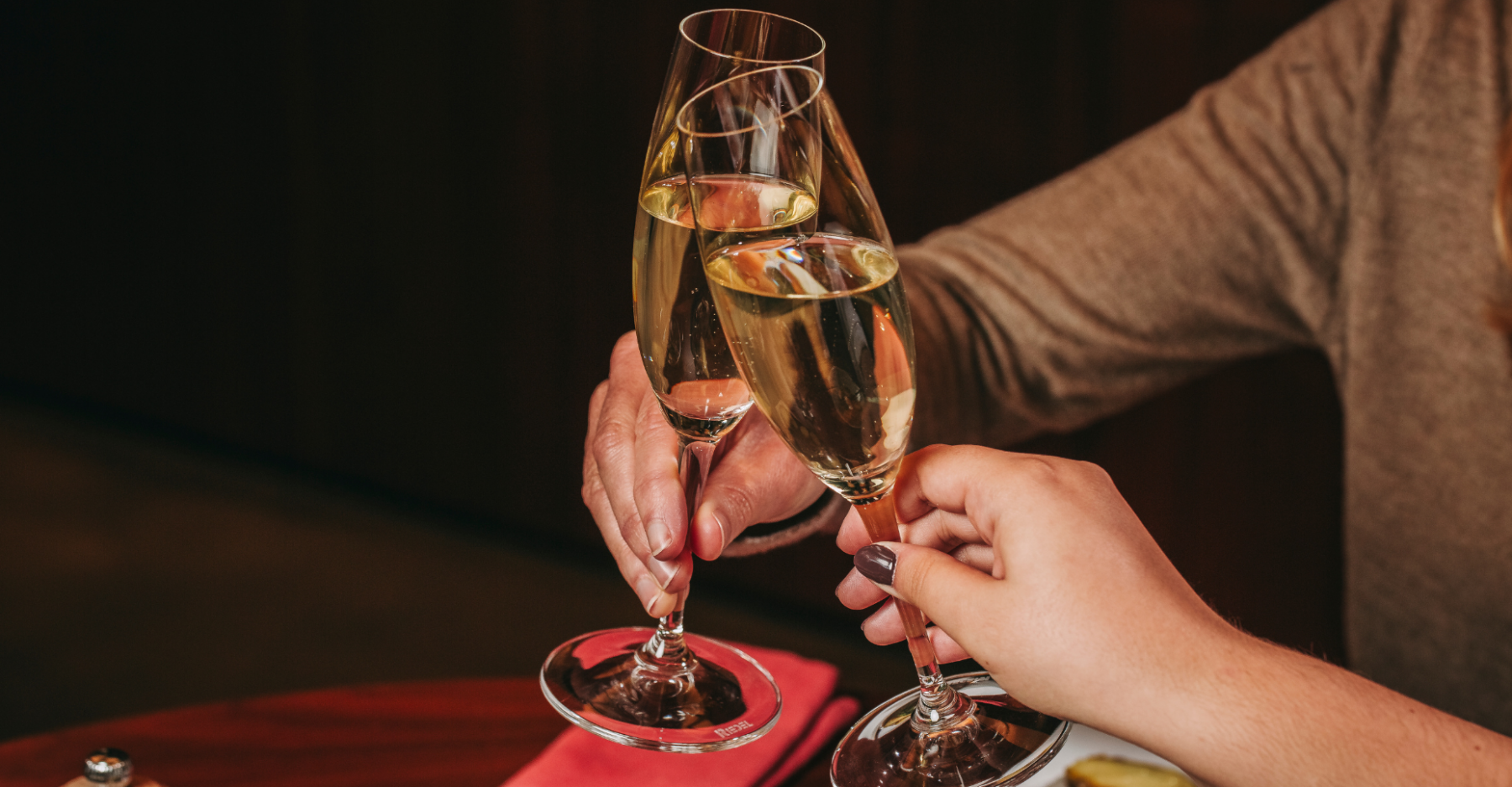 Two people clinking glasses of champagne at a table, one person wearing a brown long-sleeve top. The scene suggests a celebratory or intimate moment.