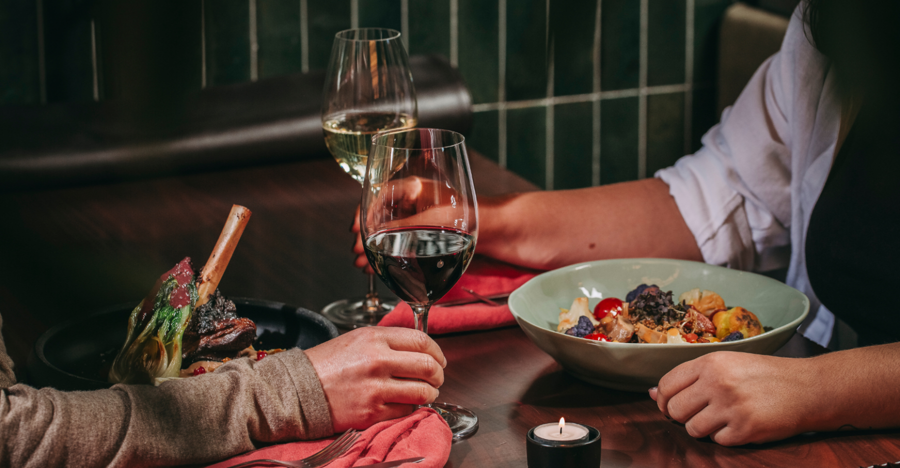Two people dining at a restaurant in Hotel Porto, holding hands across a table with wine glasses, a candle, and plates of gourmet food, including a meat dish and a colorful salad.