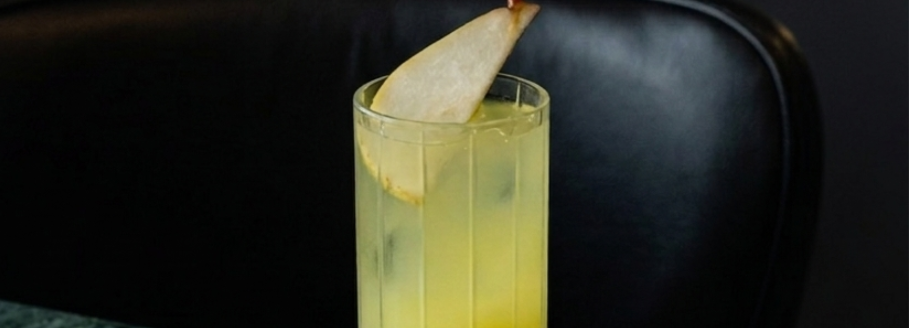 A tall glass filled with a yellow drink and ice, garnished with a thin slice of pear, sits on a dark surface in front of a black chair.