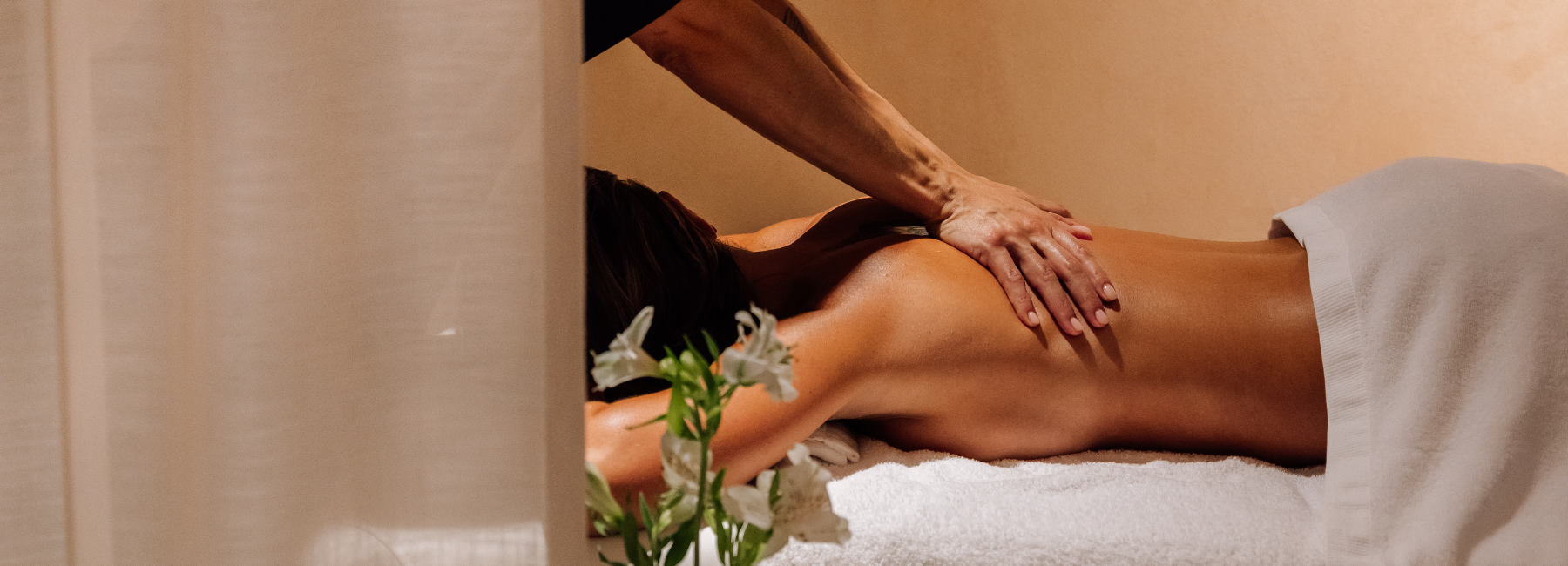 Una persona se tumba boca abajo en una camilla de masaje, cubierta con una toalla blanca, mientras otra le masajea la espalda. Una iluminación suave y flores blancas crean un ambiente de spa tranquilo y relajante.