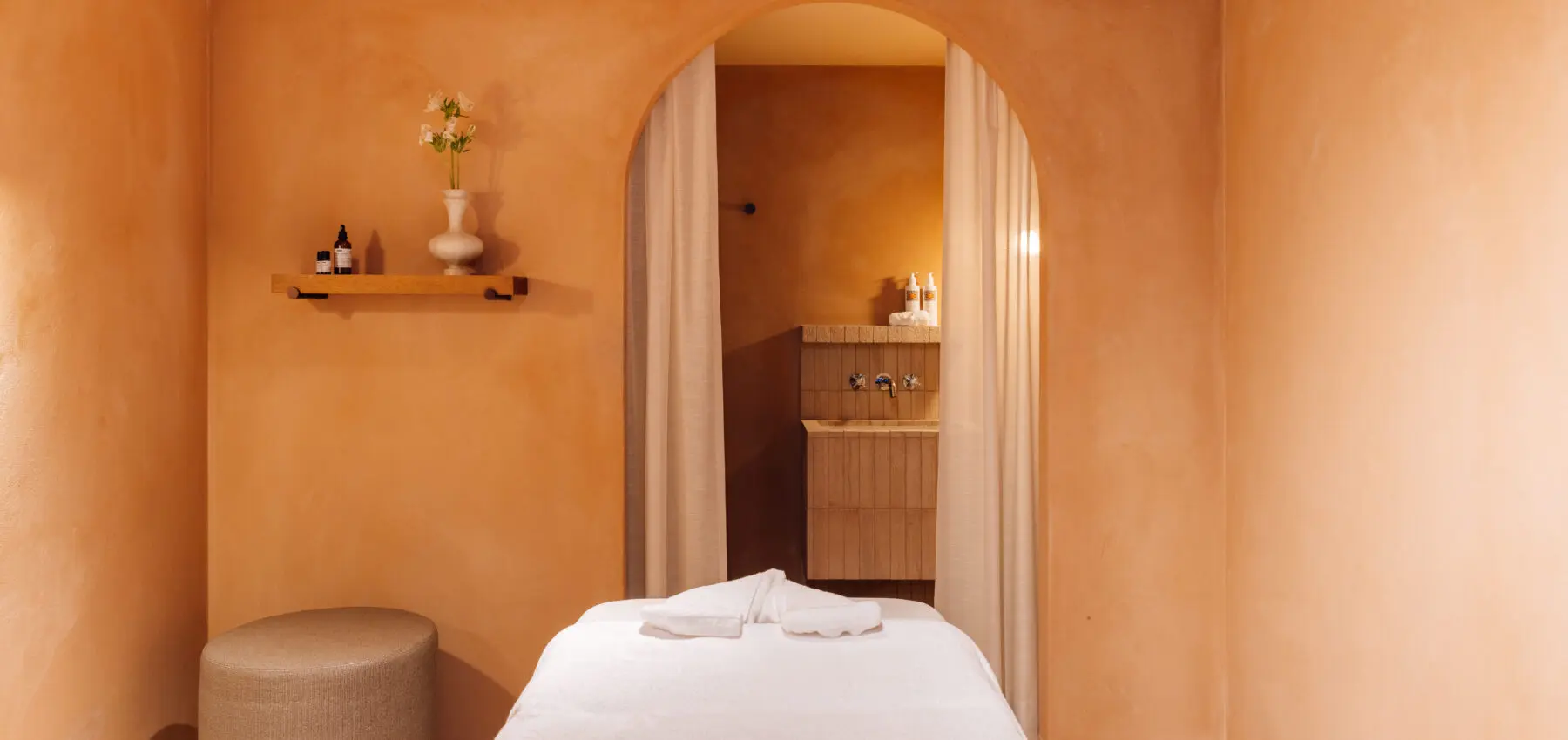 Une salle de spa sereine avec des murs couleur pêche, une table de massage recouverte de serviettes blanches, un ottoman rond, une étagère murale contenant un vase et des bouteilles, et une porte voûtée menant à un coin lavabo avec des rideaux.
