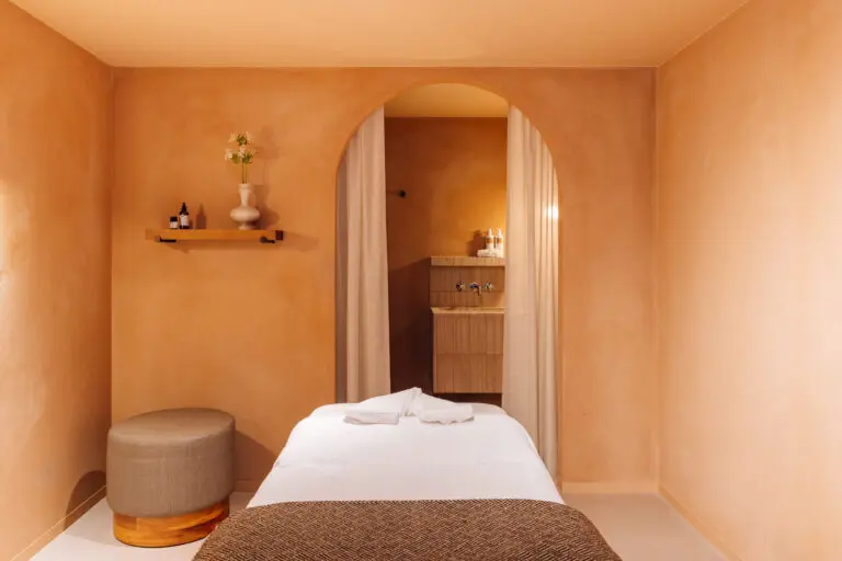 Una serena sala de spa con paredes color melocotón, una camilla de masaje cubierta de sábanas blancas, una pequeña otomana redonda, una estantería con un jarrón y botellas, y una puerta arqueada que conduce a una zona de lavabos.