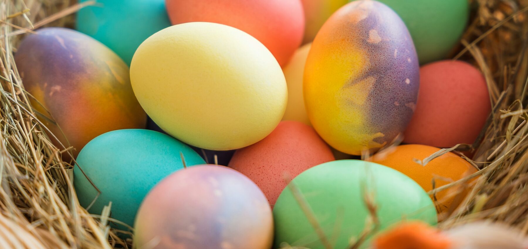 A woven basket filled with brightly coloured Easter eggs, resting on a bed of straw, with a small decorative bird perched on the edge of the basket.