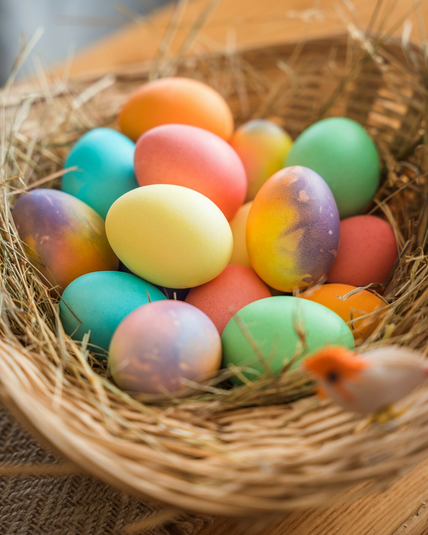 A woven basket filled with brightly coloured Easter eggs, resting on a bed of straw, with a small decorative bird perched on the edge of the basket.