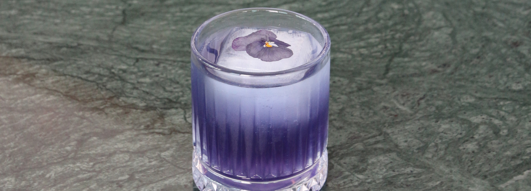 A clear glass with a purple gradient drink and a large ice sphere, topped with an edible purple flower, sits on a textured green surface.