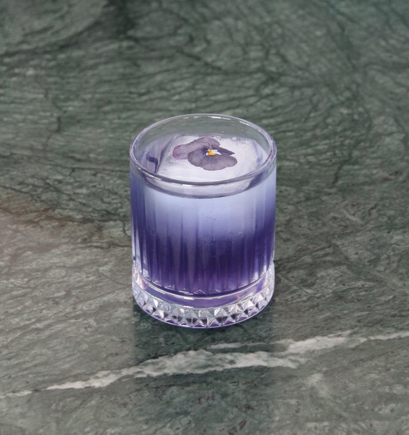 A clear glass filled with a gradient purple cocktail, topped with ice and a small purple flower, sits on a green marble surface.