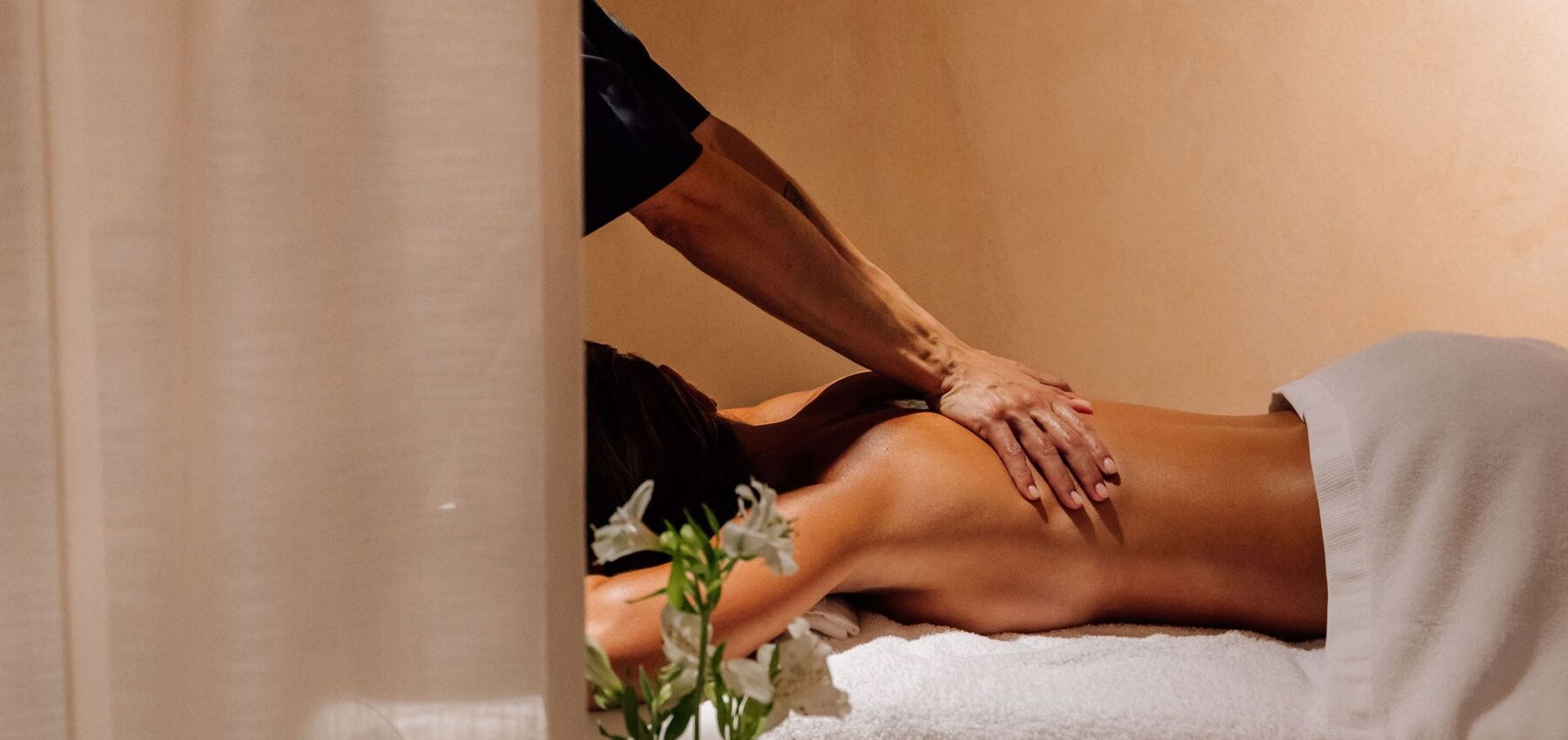 A person lies face down on a massage table, covered with a towel, while a massage therapist massages their back. A small table nearby holds a vase with flowers and a tea set. The scene is calm and softly lit.