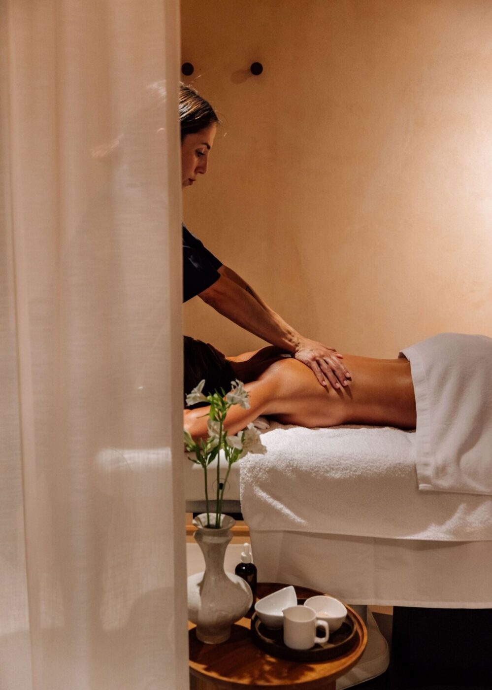 A person lies face down on a massage table, covered with a towel, while a massage therapist massages their back. A small table nearby holds a vase with flowers and a tea set. The scene is calm and softly lit.