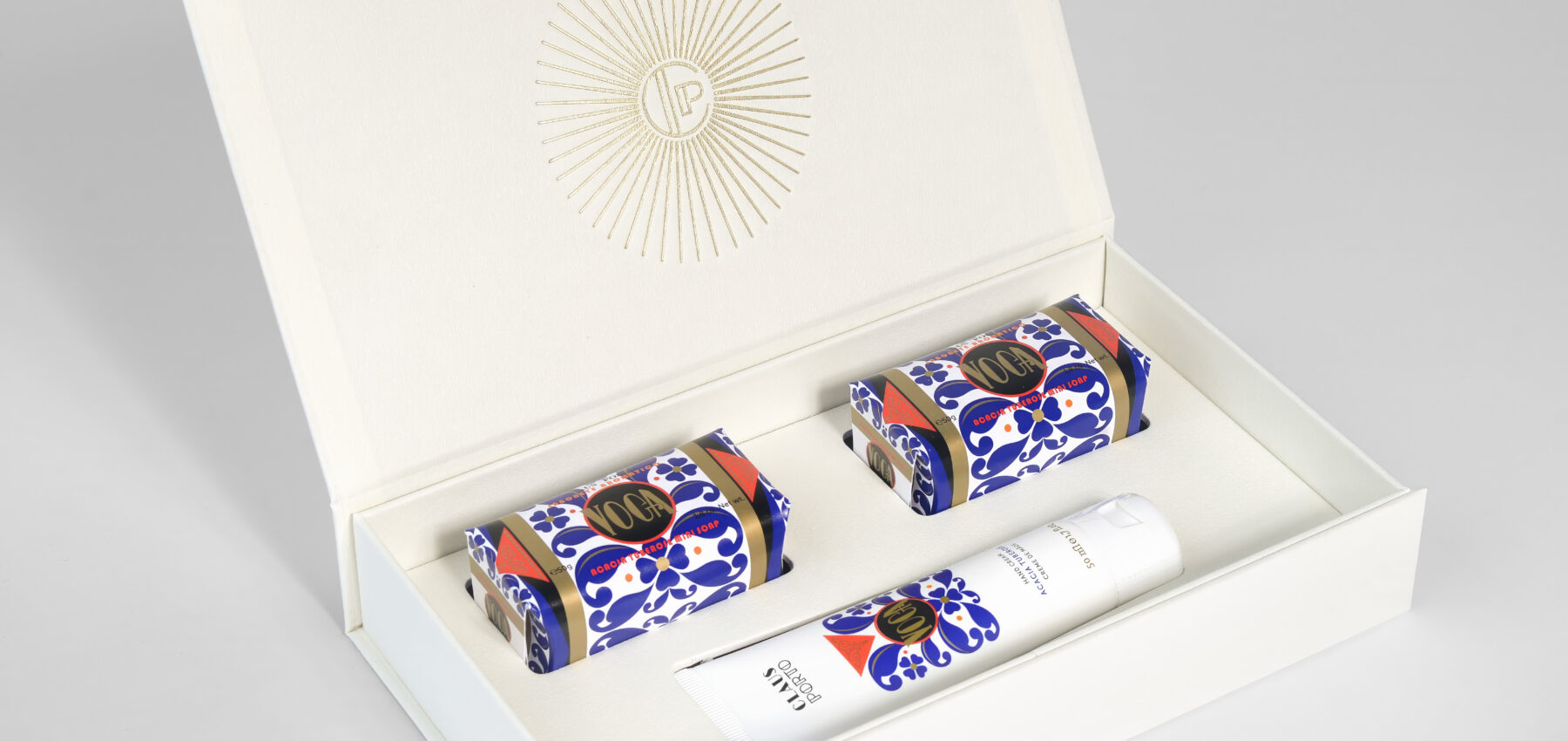 An open white gift box displays two patterned soap bars and a matching white bottle inside, all arranged neatly on a white surface. The box lid features a gold sunburst design with initials in the centre.