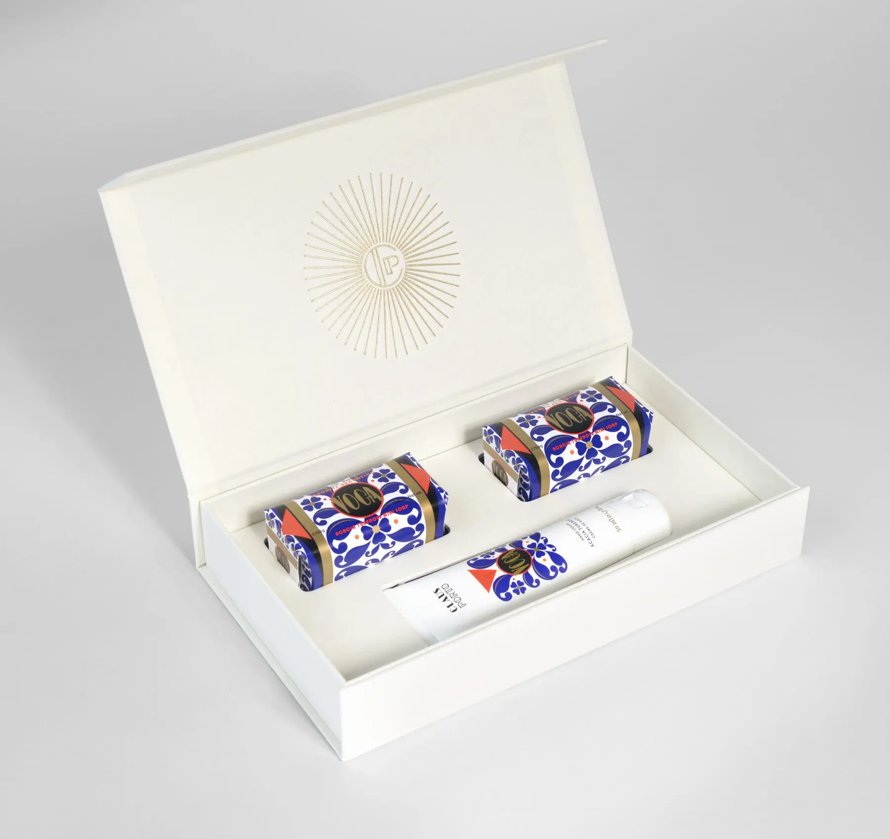 An open white gift box displays two patterned soap bars and a matching white bottle inside, all arranged neatly on a white surface. The box lid features a gold sunburst design with initials in the centre.