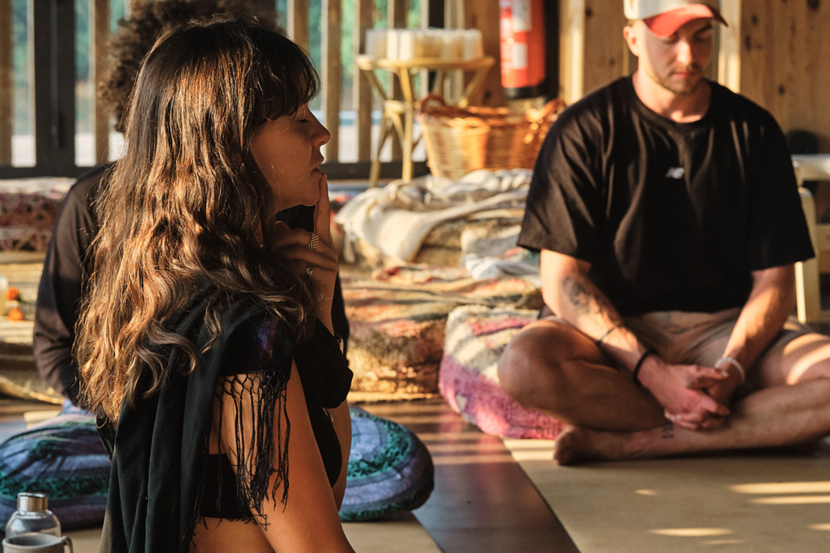 A group of people sit cross-legged on mats indoors, engaging in a meditative or mindful activity. Soft sunlight streams in, creating a calm, relaxed atmosphere. Cushions and blankets are spread around the wooden floor.