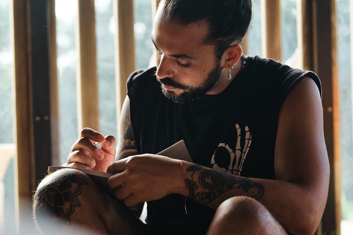 A man with a beard and tattoos sits indoors, writing in a notebook. He is wearing a black sleeveless shirt and has his hair tied back, focused on his work with sunlight streaming through wooden slats behind him.