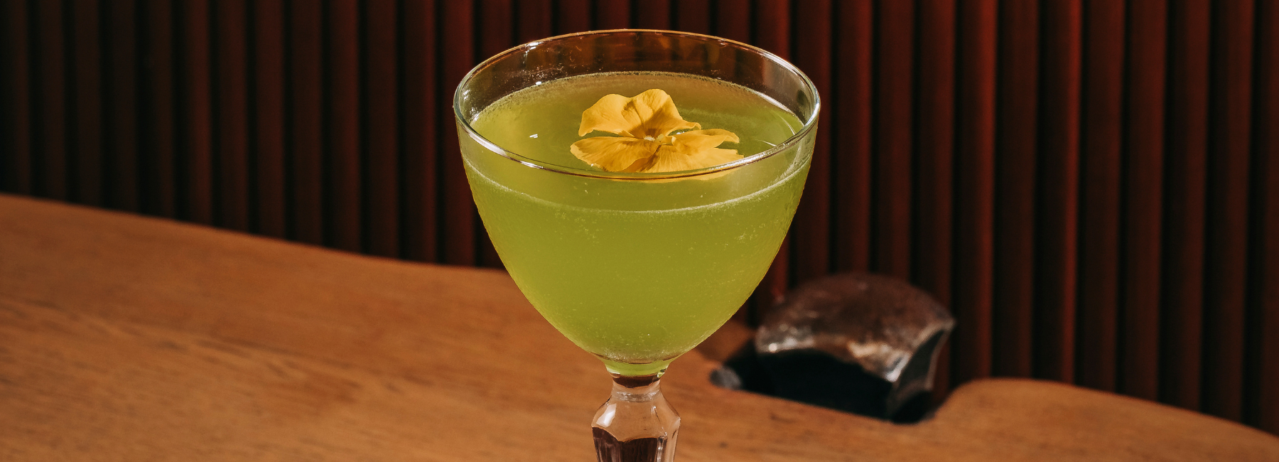 A vibrant green cocktail in a stemmed glass, garnished with a yellow edible flower, sits on a wooden surface with a dark, ribbed background.