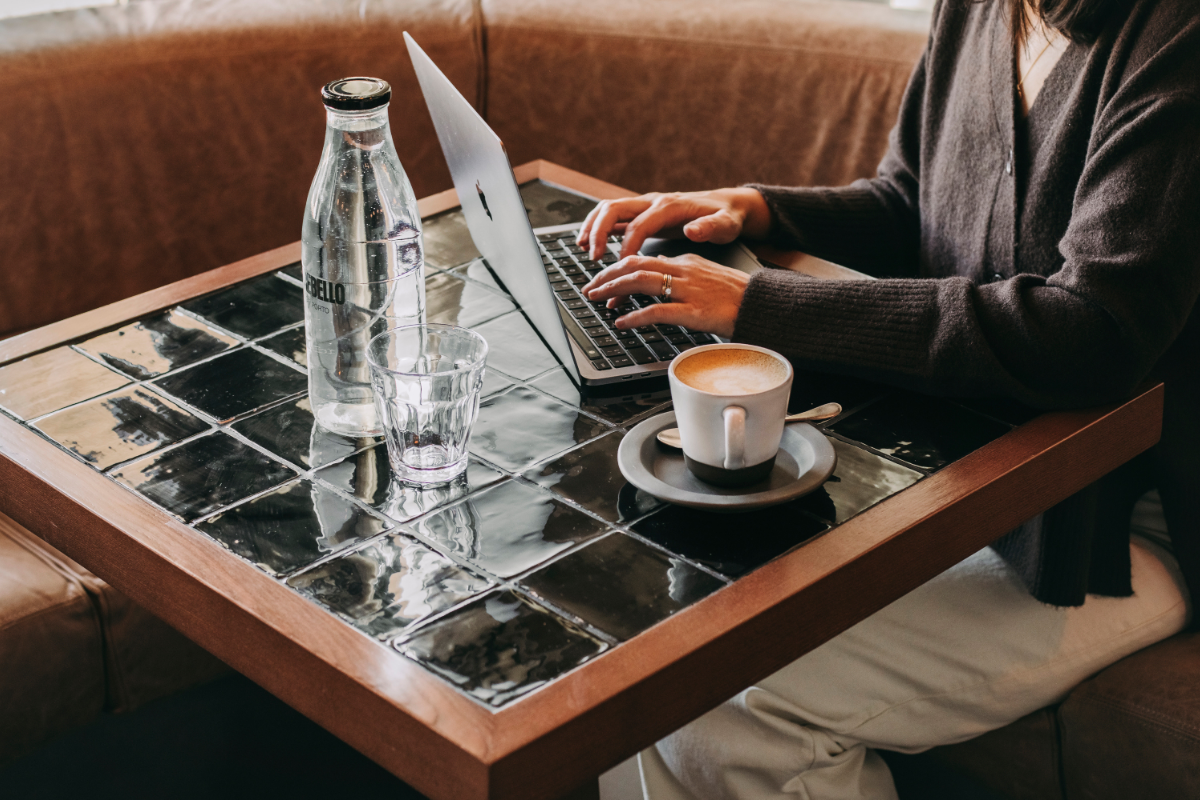 A person sits at a tiled table in a café, typing on a laptop. On the table are a glass bottle of water, an empty glass, and a cup of coffee on a saucer. The person wears a dark jumper and light trousers.