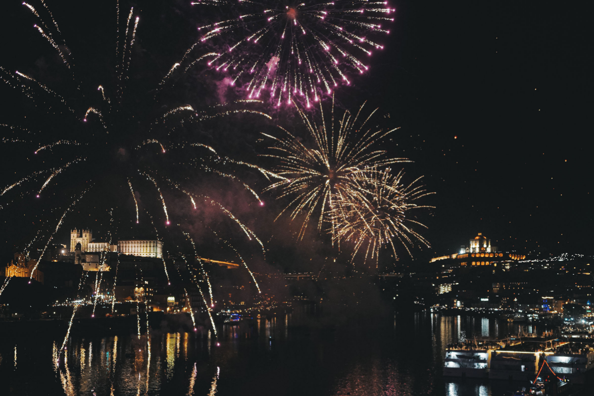 Colourful fireworks burst over a nighttime cityscape, reflecting on a river. Illuminated buildings and landmarks are visible in the background, with bright lights dotting the dark sky and water.