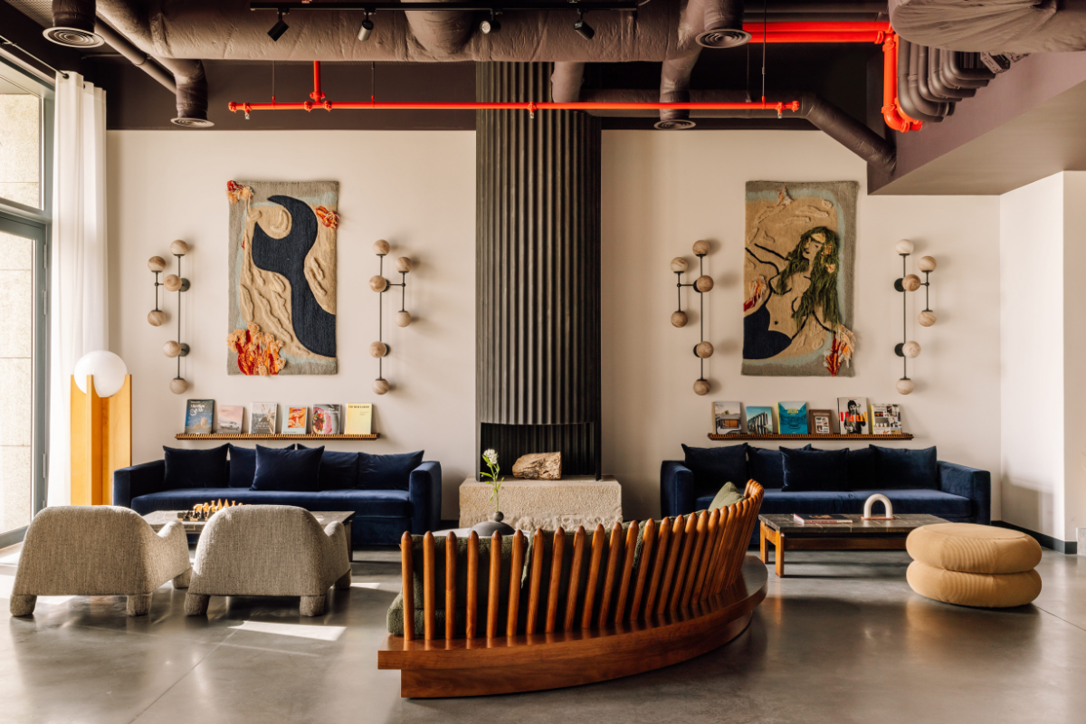 A modern lounge with velvet settees, abstract wall art, sculptural chairs, a chess set, books on display, and exposed ceiling pipes; warm lighting and neutral tones create an inviting ambience.