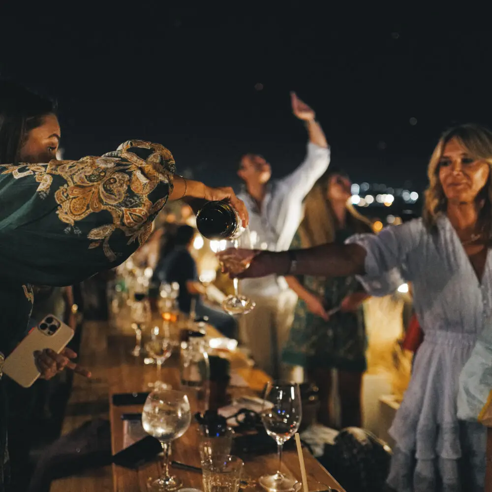 A woman in a floral sleeve pours red wine into a glass at a lively outdoor nighttime gathering, where guests savour food and wine port around a wooden table brimming with dishes, against the backdrop of shimmering city lights.