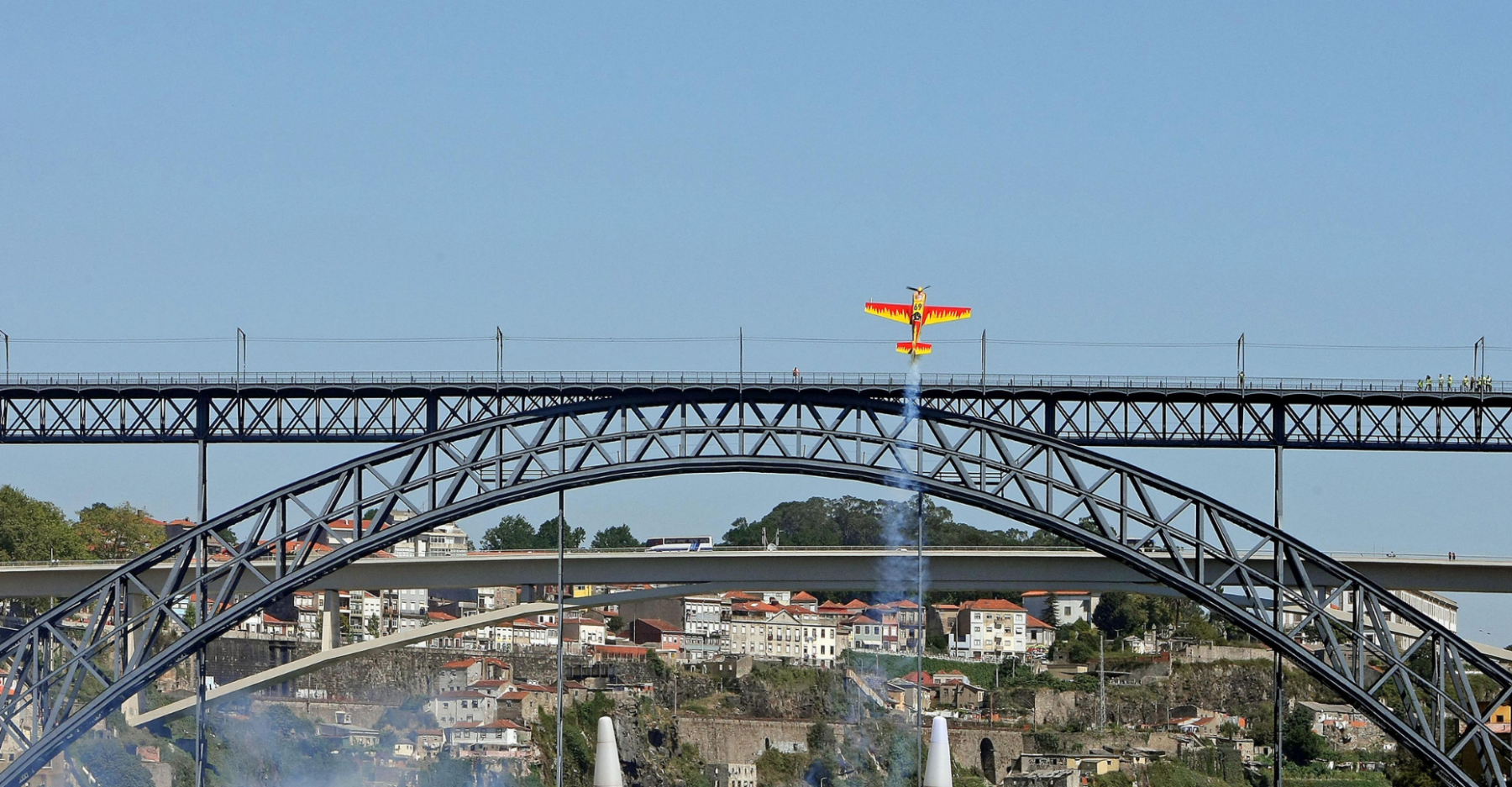 A small yellow aeroplane flies vertically, trailing smoke, above an iron bridge with an arched structure, with a cityscape and blue sky in the background.