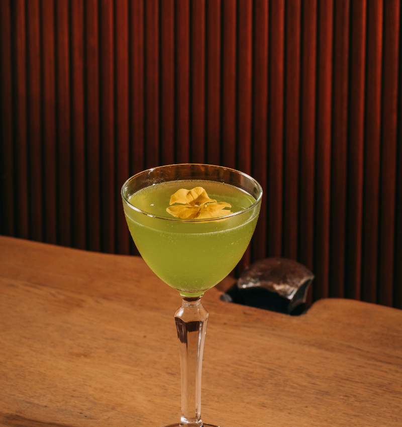 A green cocktail served in a coupe glass, garnished with a yellow flower, placed on a wooden surface with a dark, vertically-striped background.