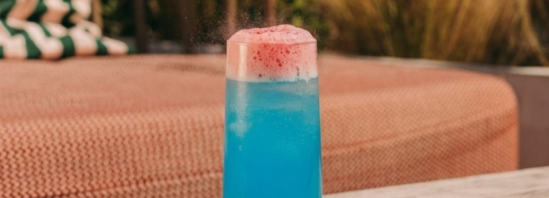 A tall glass filled with bright blue liquid is topped with a layer of pink foam. The drink sits on a table outdoors, with blurred cushions and plants in the background.