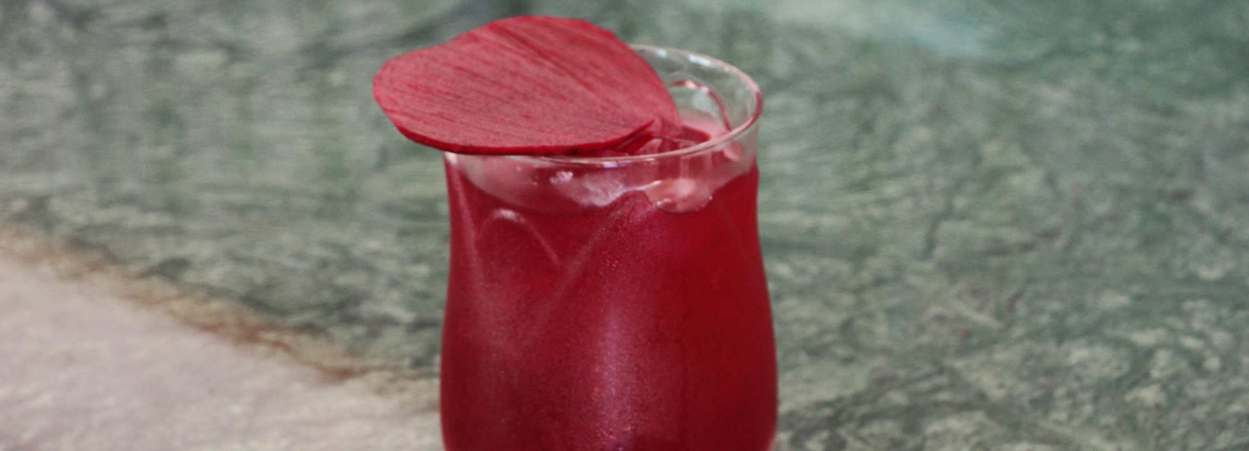 A glass of bright red drink with ice, garnished with a single red petal, sits on a textured greenish-grey surface.